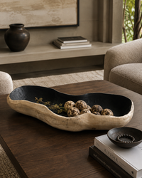 A rustic, organic-shaped wooden bowl with a black interior, styled on a dark wood coffee table in a modern living room setting. The bowl contains decorative ceramic quail eggs and features gold painted details along the rim.
