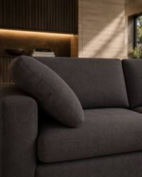 A close-up of a dark gray textured fabric sofa with a matching throw pillow, set in a modern living room with warm ambient lighting.
