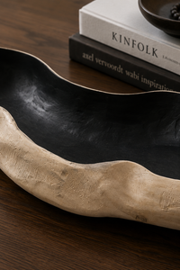 A rustic, organic-shaped serving bowl with a textured beige wood-grain exterior and a glossy black interior, styled on a dark wooden table next to books.