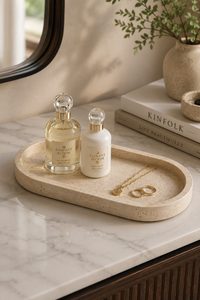 Two elegant cosmetic bottles from the brand Valtari Rose, one with a clear glass body showing pale yellow liquid and the other with a white body, both featuring gold caps and labels. They are arranged on a round beige stone tray on a white marble countertop, alongside gold jewelry and books.