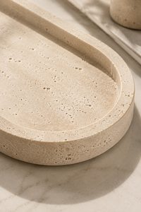 A close-up of a round travertine stone tray with a textured natural stone surface and a recessed edge, resting on a light marble surface.