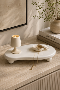 A cream-colored organic-shaped ceramic tray with small feet, styled on a light wood console table. The tray holds a small matching candle holder, a small dish with jewelry, and a gold necklace.
