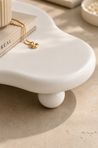 A close-up of a glossy white sculptural side table with rounded legs and a curved top, styled with a gold chain necklace draped over stacked books on a beige stone surface.