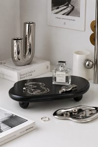 A black oval footed jewelry stand holding a silver chain necklace and a silver pocket watch, styled on a white tabletop with books, metallic vases, and a perfume bottle in the background.
