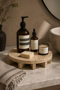 A styled bathroom scene with Aesop bath and body products arranged on a round stone tray: an amber pump bottle, an amber dropper bottle, a candle jar, and a bar of soap, set on a marble countertop beside a folded towel and a vessel with branches.