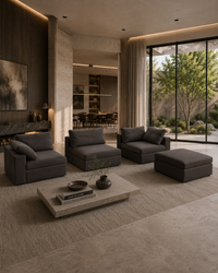 A modern living room featuring a dark gray modular sectional sofa set with matching ottomans, arranged around a low concrete coffee table on a textured rug. Large floor-to-ceiling windows offer a view of a landscaped garden.