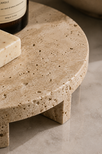 A close-up of a round travertine stone riser or tray with a speckled beige surface and blocky legs, styled with a candle and soap on a light stone floor.