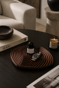 A styled interior scene showing a dark round wooden tray with concentric wave-like grooves, holding a small black bottle and a silver watch. The tray sits on a dark table beside a book, a bowl, a lit candle, and part of a sofa in a real home environment.