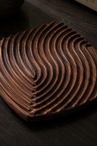 A dark brown wooden tray with a wavy, layered design, photographed on a dark wood surface.