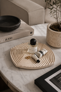 A beige carved wooden tray with concentric wave patterns sits on a white marble table, styled with a small perfume bottle, a tube of body cream, and a gold ring. The scene is set in a modern living room with a beige sofa, a potted plant, and a book in the background.