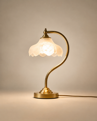 A brass table lamp with a glowing white flower-shaped glass shade, mounted on a curved brass stem with a round base, set against a warm beige background.