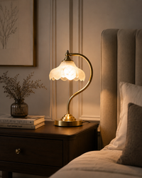 A warm brass bedside table lamp with a glowing white scalloped glass shade, styled on a dark wood nightstand next to a bed in a cozy bedroom.