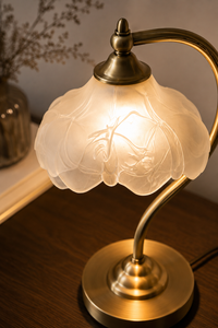 A vintage-style table lamp with a warm brass-colored metal base and curved arm, featuring a frosted glass shade with an embossed floral pattern that glows with a soft yellow light. The lamp is shown turned on on a dark wooden surface with a blurred background.