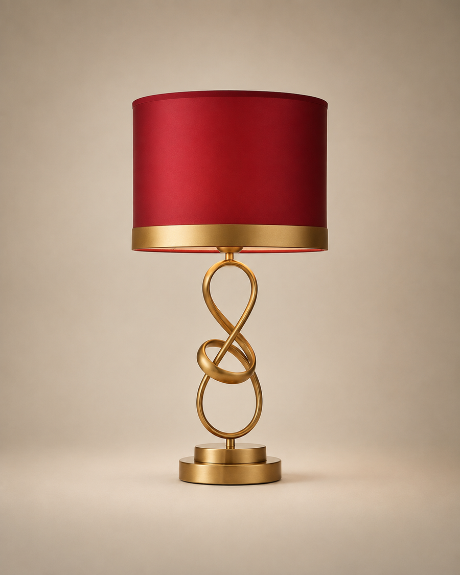 A table lamp with a cylindrical red fabric shade, a gold metallic band at the base of the shade, and an ornate gold metal sculptural base on a warm beige background.