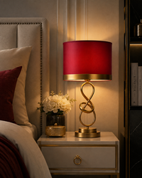 A modern table lamp with a red fabric drum shade and a gold-finished sculptural base, styled on a bedside table next to a bed in a warm, elegant bedroom setting.