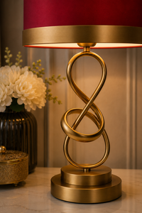 A modern table lamp with a gold-finished sculptural infinity symbol base and a deep red fabric shade, styled on a marble surface next to a vase of white flowers.
