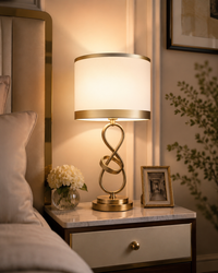 A modern table lamp with a glowing white drum shade and a gold-finished twisted infinity base, styled on a marble nightstand in a bedroom setting.