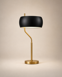 A modern table lamp with a matte black drum shade and a brushed gold metal stem and base, set against a warm beige studio background.