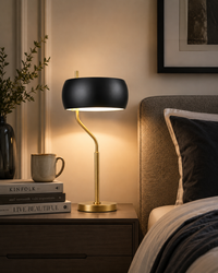 A modern table lamp with a matte black dome shade and a curved gold metal stem, illuminated on a wooden bedside table next to a bed.