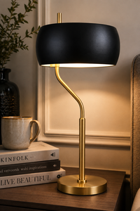 A modern table lamp with a matte black dome shade and a curved gold metal stem, illuminated on a wooden bedside table next to a mug and books.