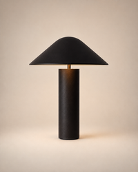 A modern table lamp with a matte black conical shade and a cylindrical black base, illuminated with a warm glow from within, set against a solid beige background.
