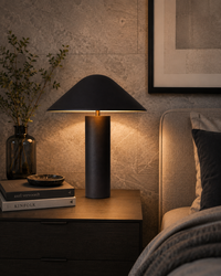 A modern table lamp with a matte black conical shade and cylindrical base sits on a dark wooden bedside table in a styled bedroom setting, casting a warm glow next to a bed and a textured wall.