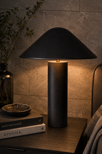 A modern table lamp with a matte black conical shade and cylindrical base, illuminated to cast a warm glow, styled on a dark wooden bedside table next to books and a vase.