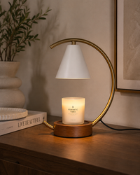 A modern table lamp with a curved gold metal frame, a white cone shade, and a warm wooden base holding a lit white candle. The lamp sits on a wooden table in a styled interior setting with a potted plant and framed wall art in the background.