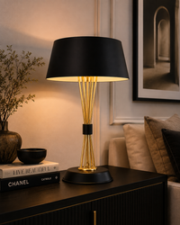 A modern table lamp with a black drum shade and a gold-finished sculptural base, lit warmly on a dark wooden sideboard in a styled interior setting.