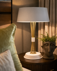 A modern table lamp with a white drum shade, a gold-finished stem made of clustered rods, and a white circular base, styled on a dark wood bedside table next to a potted plant.