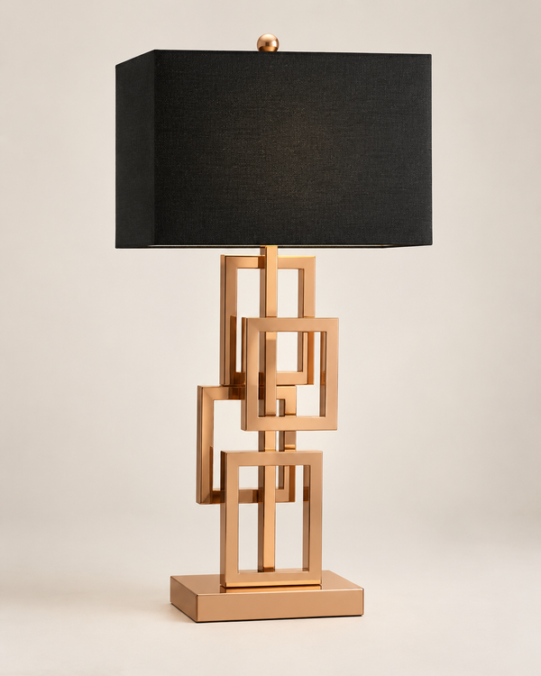 A modern table lamp with a black rectangular fabric shade and a metallic rose gold base made of interlocking geometric square frames, set against a plain light beige background.