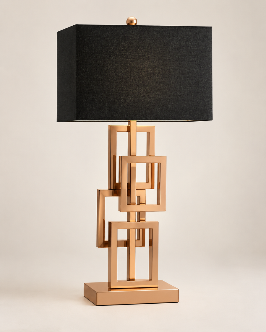 A modern table lamp with a black rectangular fabric shade and a metallic rose gold base made of interlocking geometric square frames, set against a plain light beige background.