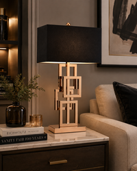 A modern table lamp with a black rectangular fabric shade and a geometric gold metal base, styled on a marble-topped side table in a cozy living room setting.
