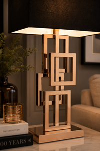 A modern table lamp with a geometric rose gold metal frame and a black fabric drum shade, shown lit on a tabletop in a styled interior setting.