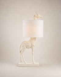 A white giraffe-shaped table lamp with a cylindrical white shade, standing on a rectangular base against a neutral beige background.