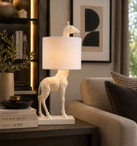 A sculptural giraffe table lamp with a white geometric textured body and a white drum shade, lit with a warm glow, set on a dark wood side table in a modern living room next to a beige sofa.