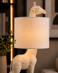 A table lamp shaped like a giraffe with a textured cream-colored body and a large cylindrical fabric shade, shown lit in a warm indoor setting.
