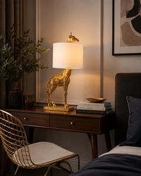 A lit table lamp with a sculptural gold horse base and a white drum shade sits on a dark wood bedside table in a styled bedroom. A gold wire chair, books, a plant, and part of a bed are visible around it.