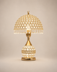 A gold-finished table lamp with a dome-shaped shade and a spherical base, both decorated with clear crystal beads and hanging crystal fringe, shown lit against a plain beige background.