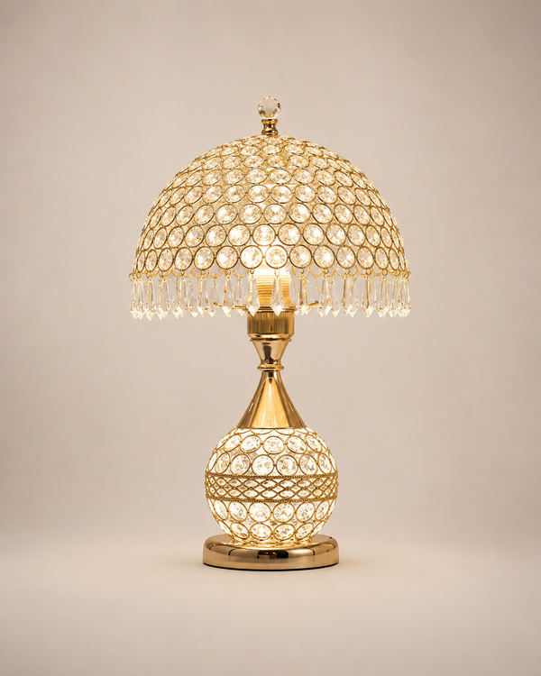 A gold-finished table lamp with a dome-shaped shade and a spherical base, both decorated with clear crystal beads and hanging crystal fringe, shown lit against a plain beige background.