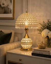 A glowing table lamp with a gold-finished base and a crystal-like dome shade, placed on a white side table in a styled living room setting with books, a vase of flowers, and a sofa visible.