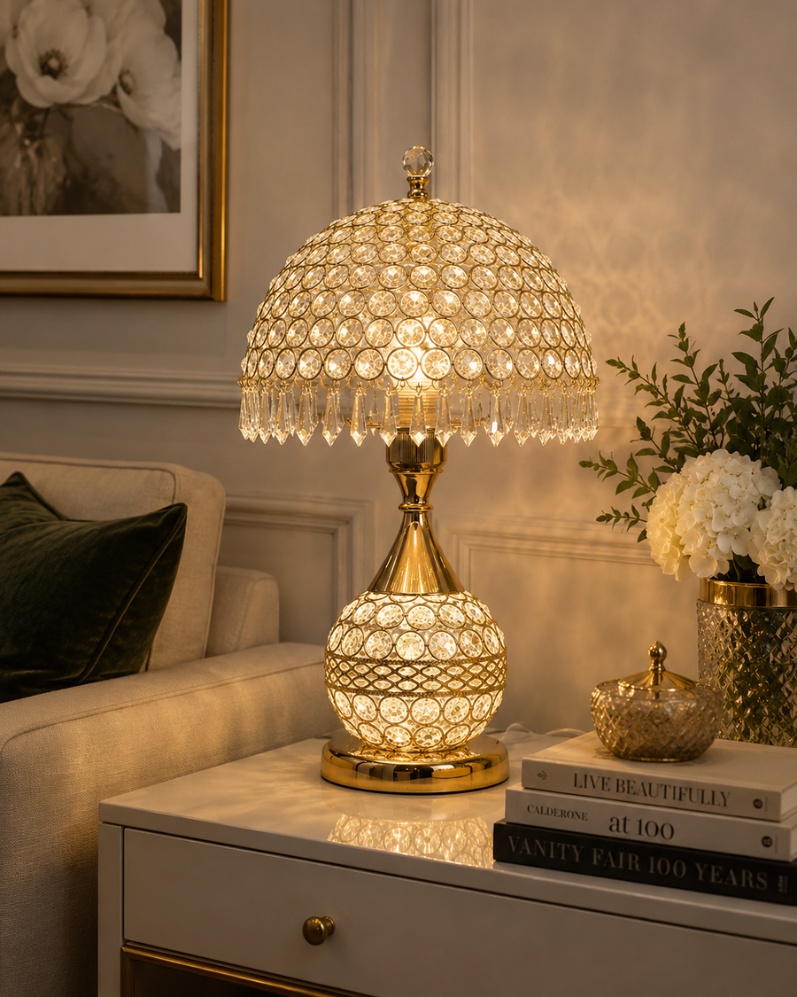 A glowing table lamp with a gold-finished base and a crystal-like dome shade, placed on a white side table in a styled living room setting with books, a vase of flowers, and a sofa visible.