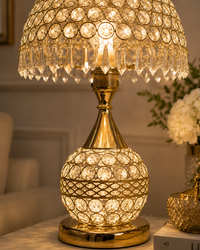 A gold table lamp with a crystal bead-covered shade and matching crystal-covered spherical base, illuminated with a warm glow in an indoor setting.