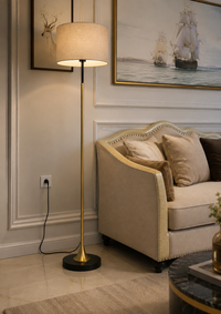 A modern floor lamp with a beige drum shade and a brushed gold metal stand is shown in a styled living room next to a beige sofa with decorative pillows.