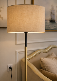 A modern floor lamp with a large beige linen drum shade, a black upper stem, and a brushed gold lower stem, shown lit in a styled interior next to a beige armchair.