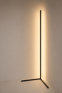 A modern black metal floor lamp with a minimalist L-shaped base and a slim vertical light bar, casting a warm yellow glow against a plain off-white wall.
