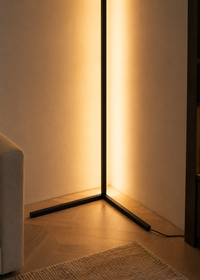 A modern black metal floor lamp with a tall vertical light panel casting a warm golden glow in a minimalist room corner with light wood flooring.