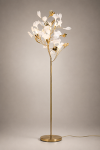 A modern tree-style floor lamp with a slim gold metal stem and circular base, topped with illuminated white flower-like petals and small gold accents, photographed against a neutral warm gray studio background.