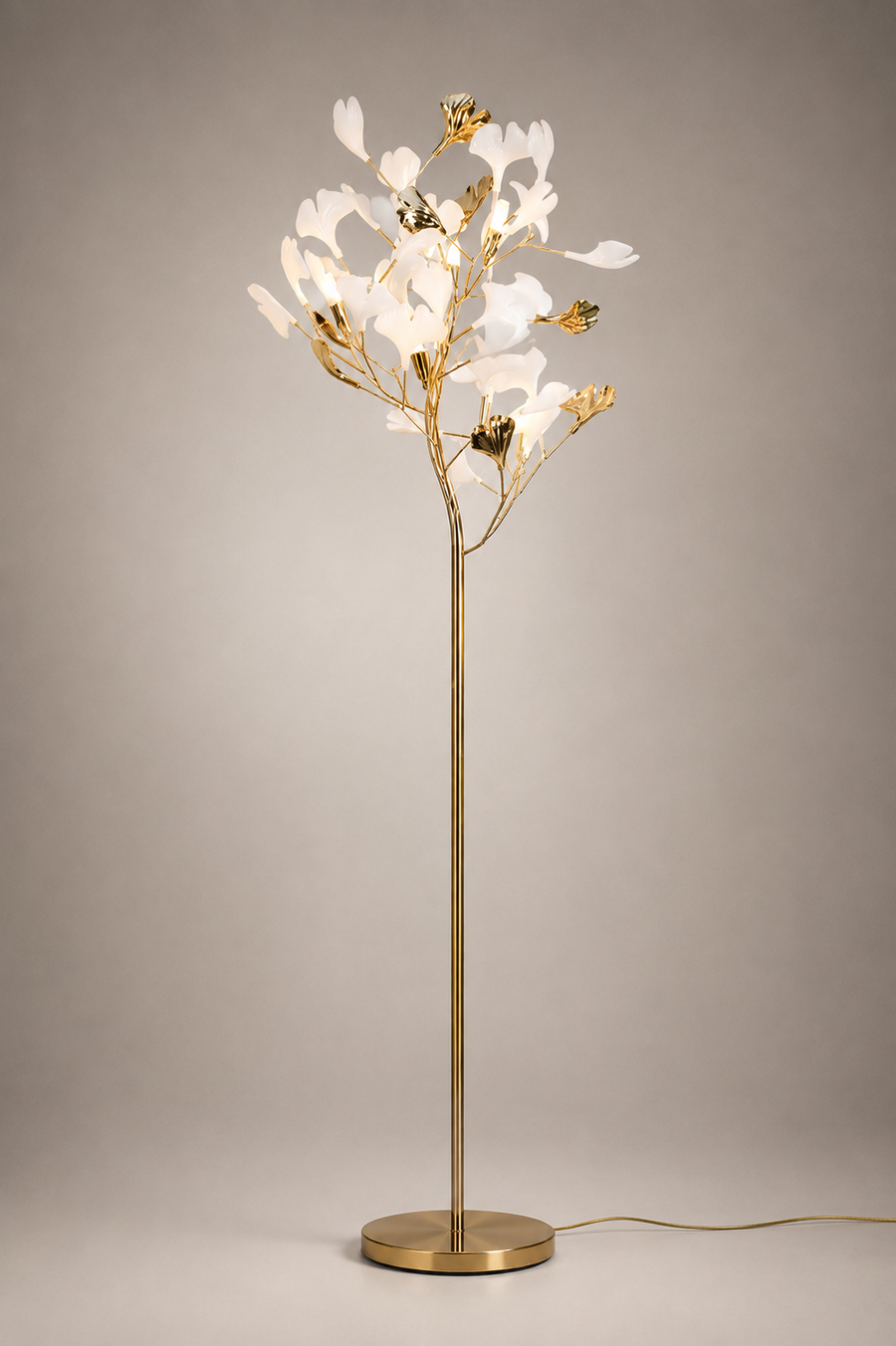A modern tree-style floor lamp with a slim gold metal stem and circular base, topped with illuminated white flower-like petals and small gold accents, photographed against a neutral warm gray studio background.
