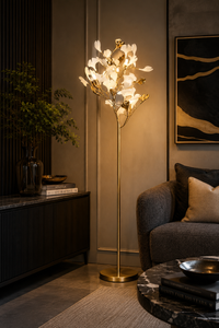 A modern floor lamp with a gold metal stand and white flower-shaped bulbs, illuminated in a warm light, placed in a stylishly decorated living room next to a gray armchair.